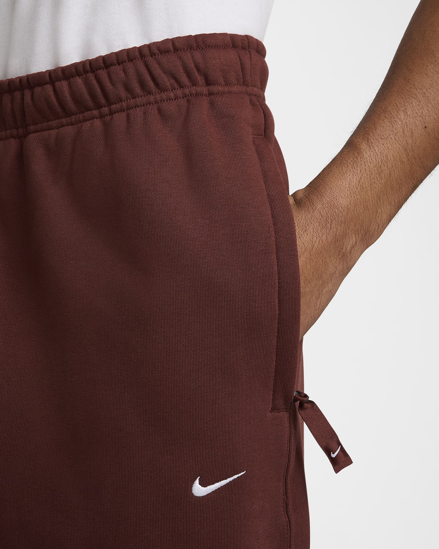 M+NK+SOLO+SWSH+FLC+CF+PANT (2) Hand in pocket of brown Nike sweatpants with logo