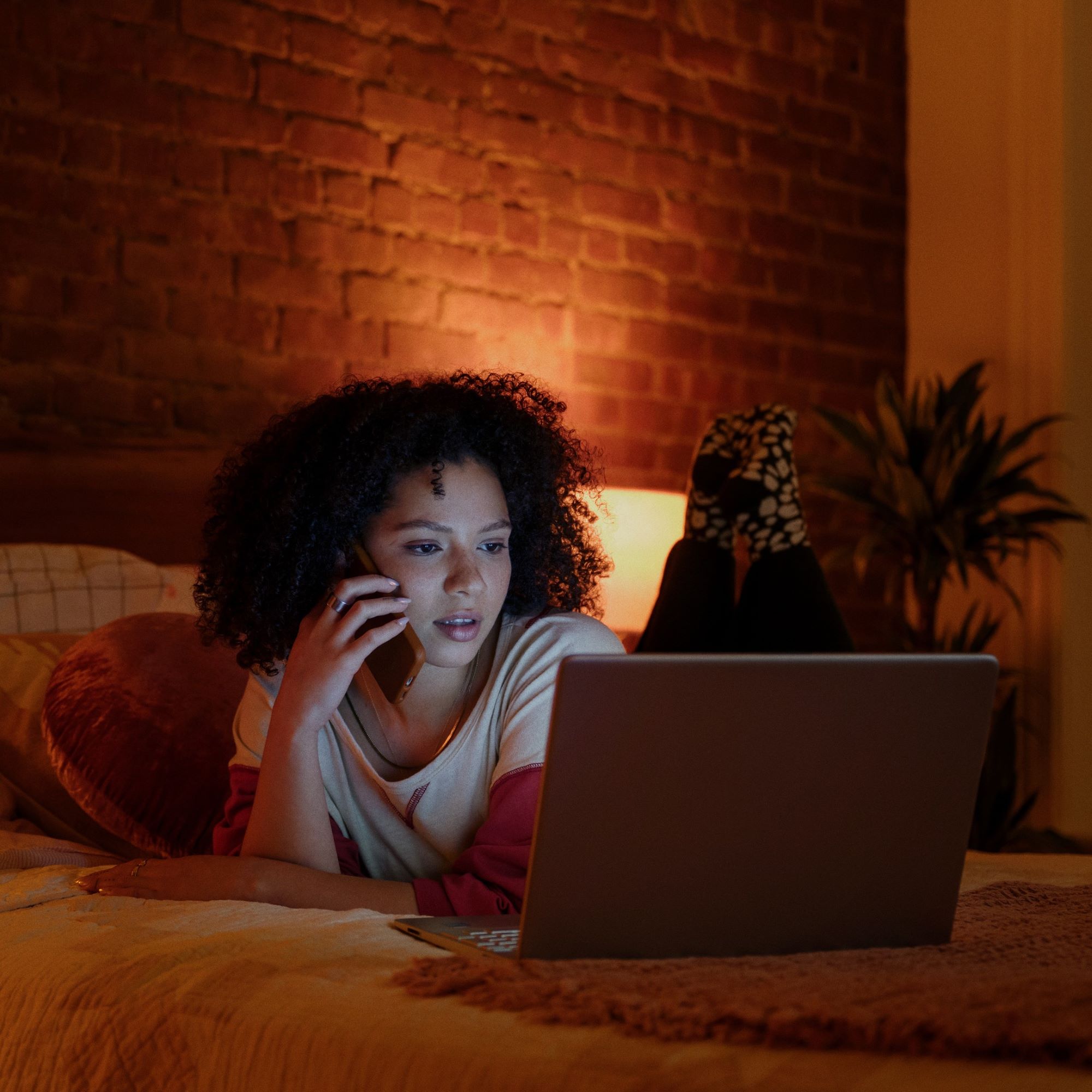 Woman lying on bed using laptop and talking phone