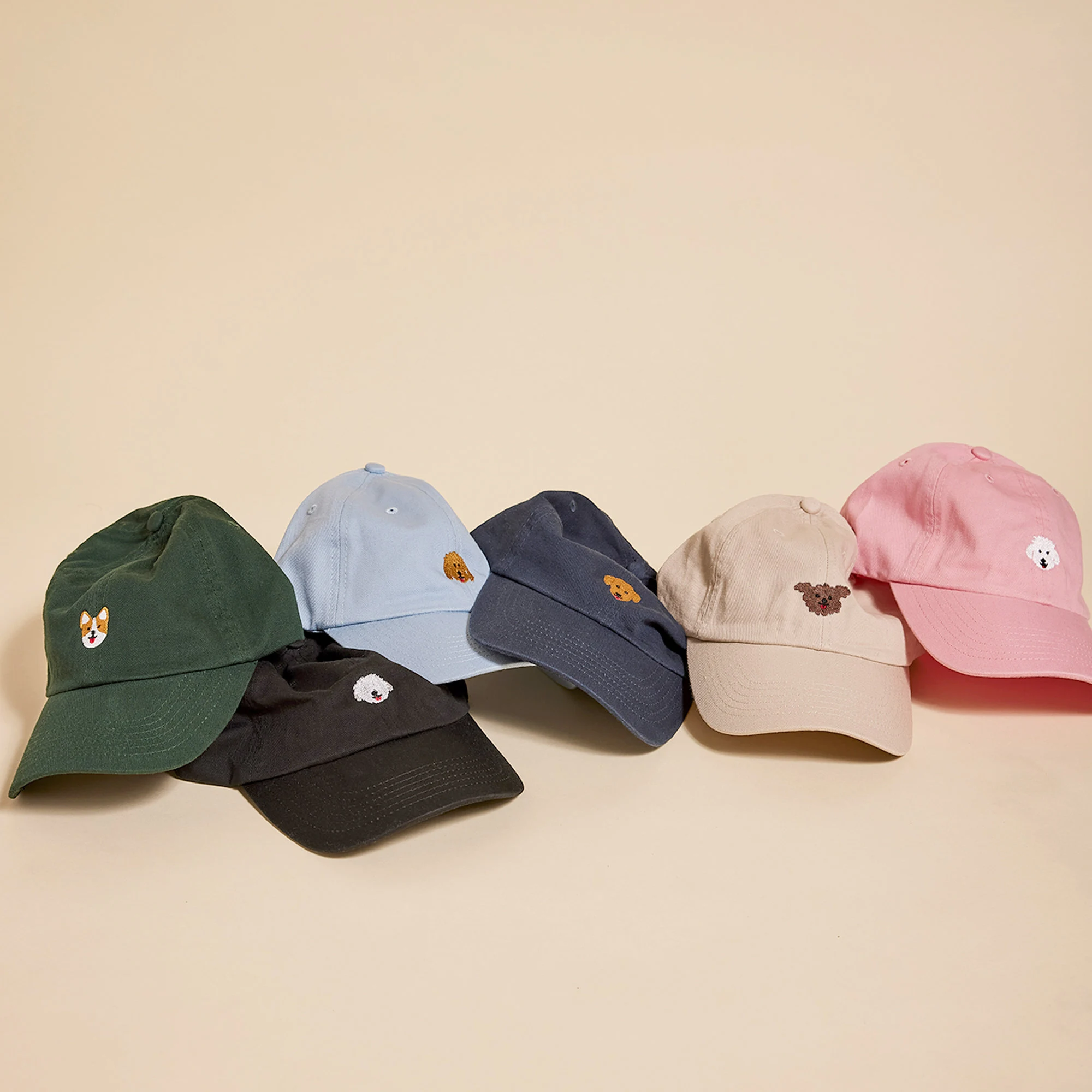 Six baseball caps in different colors arranged in a row on a beige background, each featuring a small embroidered dog face on the front, including green, black, light blue, navy, beige, and pink caps.