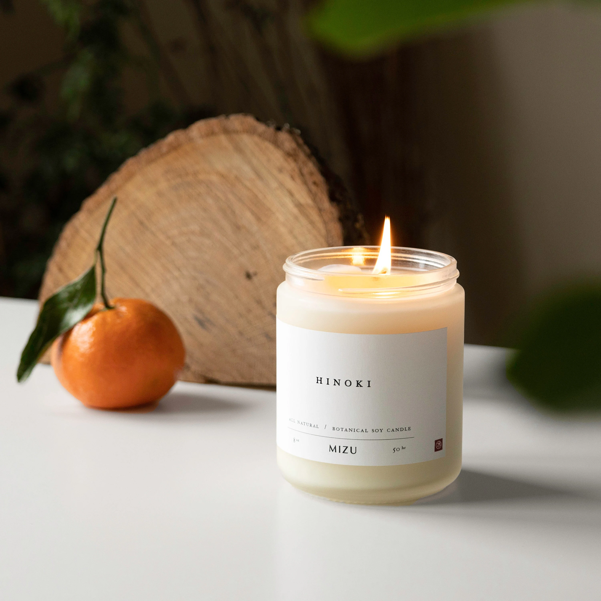 A lit Hinoki Mizu botanical soy candle in a frosted glass jar on a white surface, with a fresh mandarin orange with leaves and a wooden tree slice in the background.